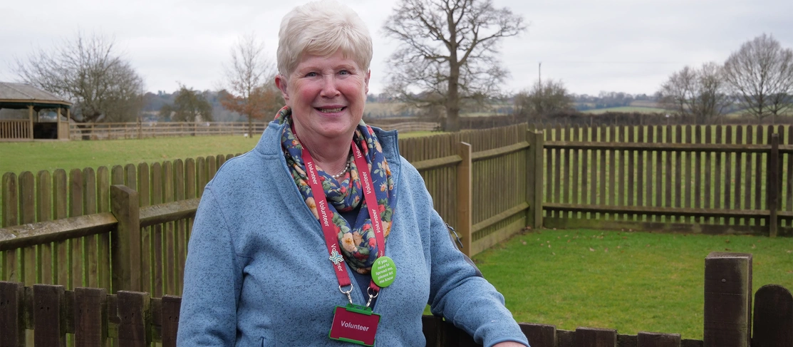 Mid shot of a happy woman leaving on a fence outside looking directly at the camera and wearing a lanyard with a badge that says ‘Volunteer’