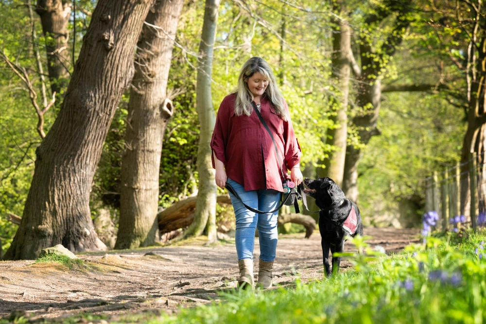 A woman walking a black labrador through a forest path. They are looking at each other and the woman is smiling.