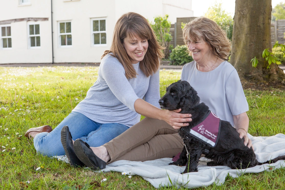 Two women sat on a blanket on some grass with a spaniel sat next to them in a hearing dogs jacket