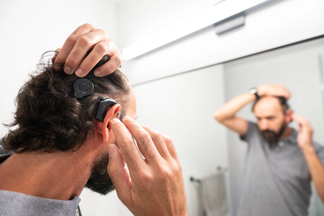 A man adjusting his cochlear implant in a room with a mirror so the back of his head and the reflection of his face is visible