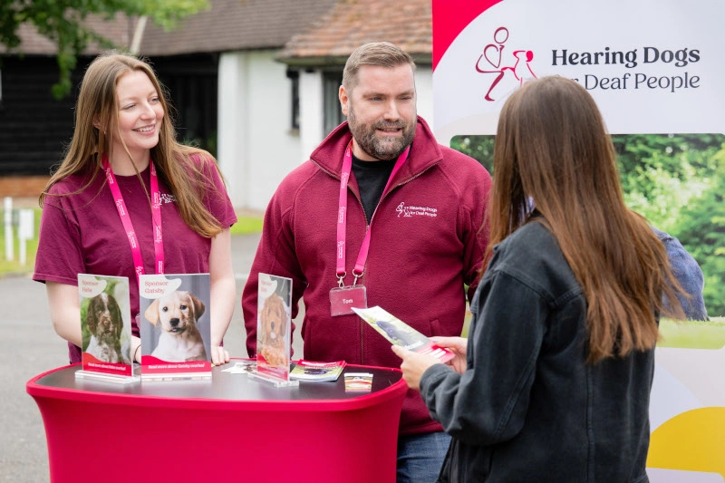 A man and 2 women are standing around a red table with brochures and pamphlets on it. Behind them is a banner that reads "Hearing Dogs for Deaf People"