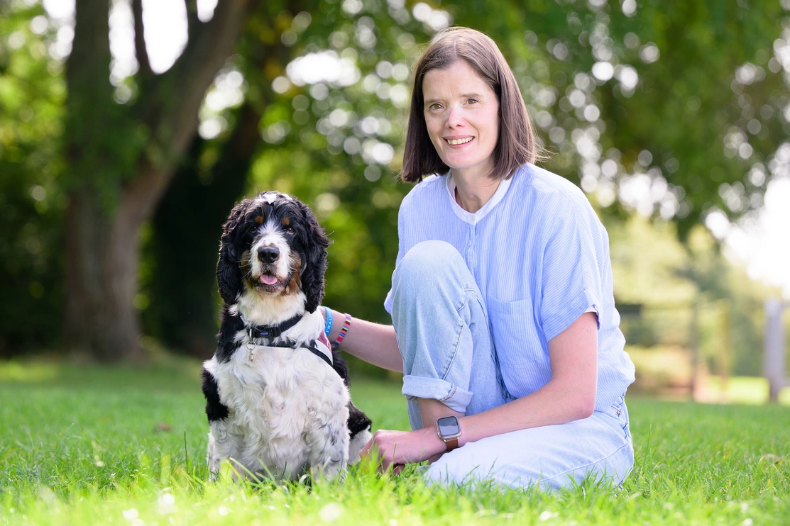 A woman is kneeling on grass beside a dog in a park-like setting. Out of focus trees can be seen in the background.