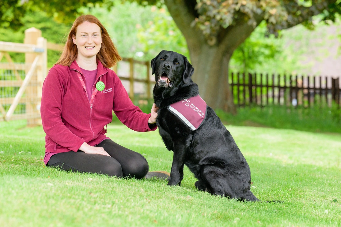 A hearing dogs member of staff sat outside with a black labrador in a hearing dogs jacket