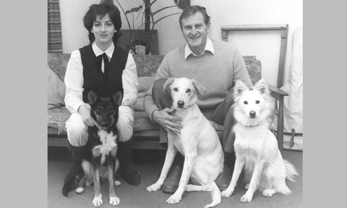 An old photo of a woman and a man sat in a lounge with three dogs sat in front of them