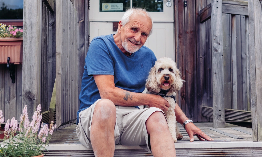 A man wearing a shorts and blue tshirt is sitting on porch steps with a white cockapoo dog