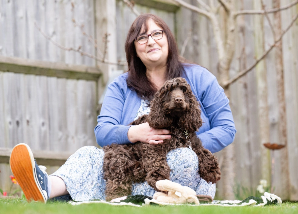 A bespectacled woman wearing a blue cardigan over a long dress is sitting on grass holding a brown curly haired dog in her arms.