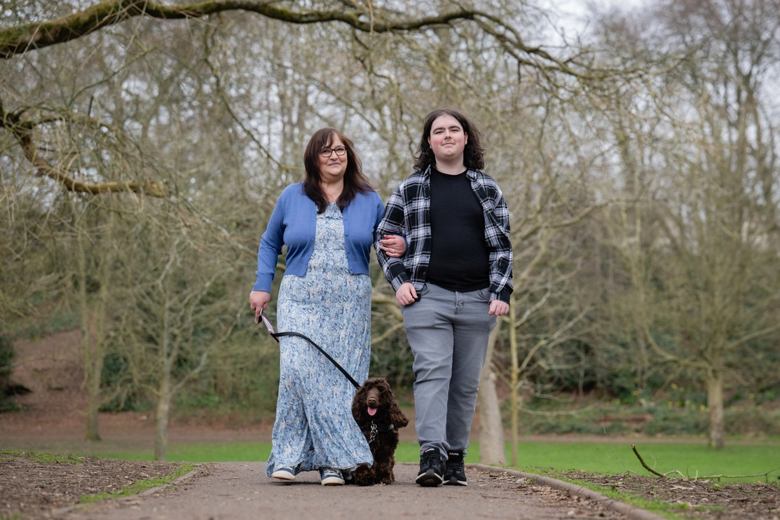 A woman wearing a blue cardigan and long dress is walking with a young man wearing a checkered shirt over black tshirt. The woman is holding the leash of a brown dog with its tongue out.