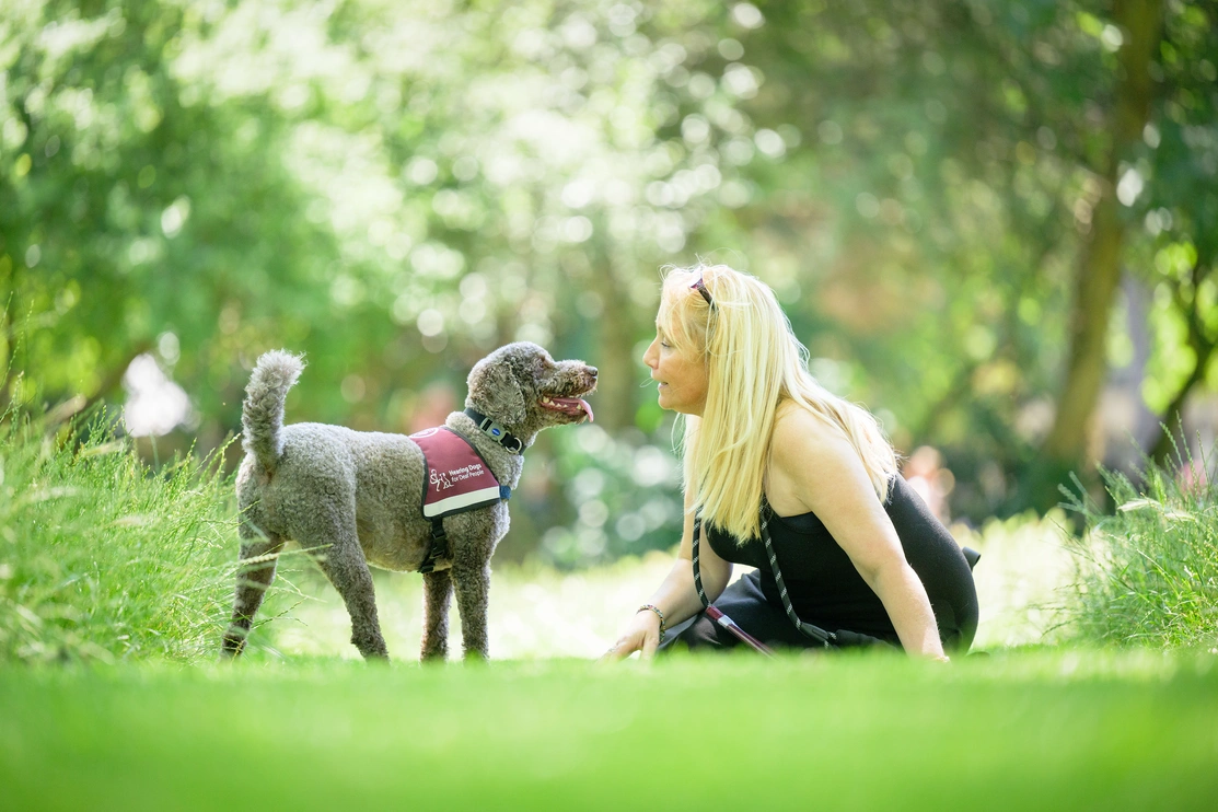 Aa blonde woman sitting on grass surrounded by trees. A hearting dog with jacket is standing in front of her