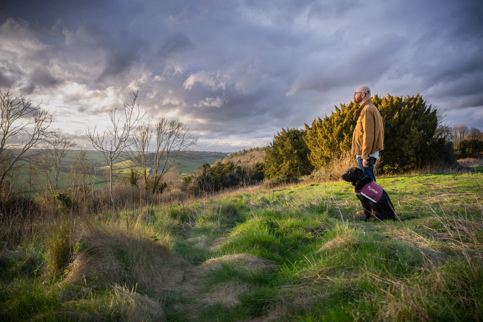 A photograph of Trevor and a black hearing dog gazing out across a dramatic outdoor scene of wild grass, green hills and clouds.