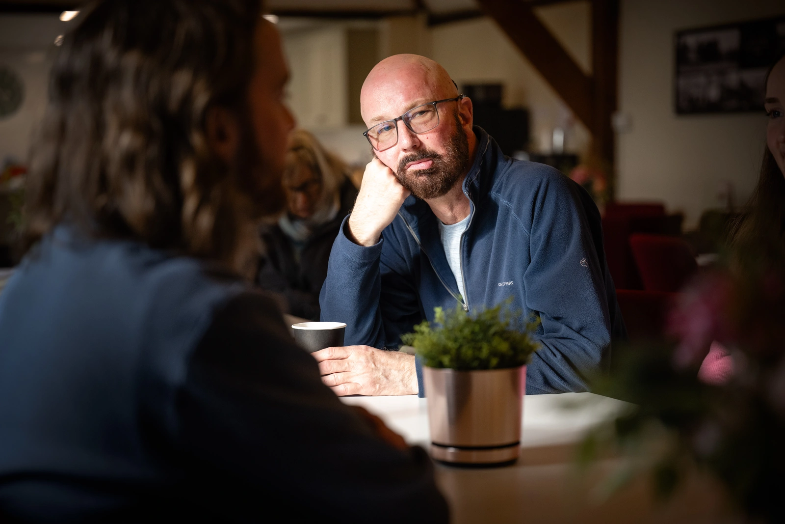 A bearded, bespectacled man sitting in a cafe. He looks sad and is resting his face on his fist.