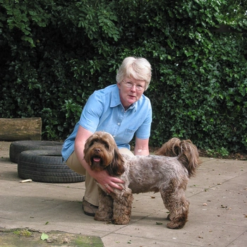 A women crouching down next to a brown cockapoo outside with a hedge behind them, both are looking at the camera
