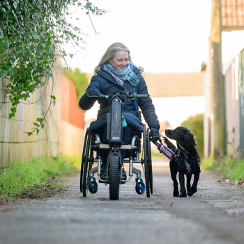 Woman in a wheelchair walking with a hearing dog on a lead down a path outside 