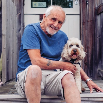 A man wearing a shorts and blue tshirt is sitting on porch steps with a white cockapoo dog