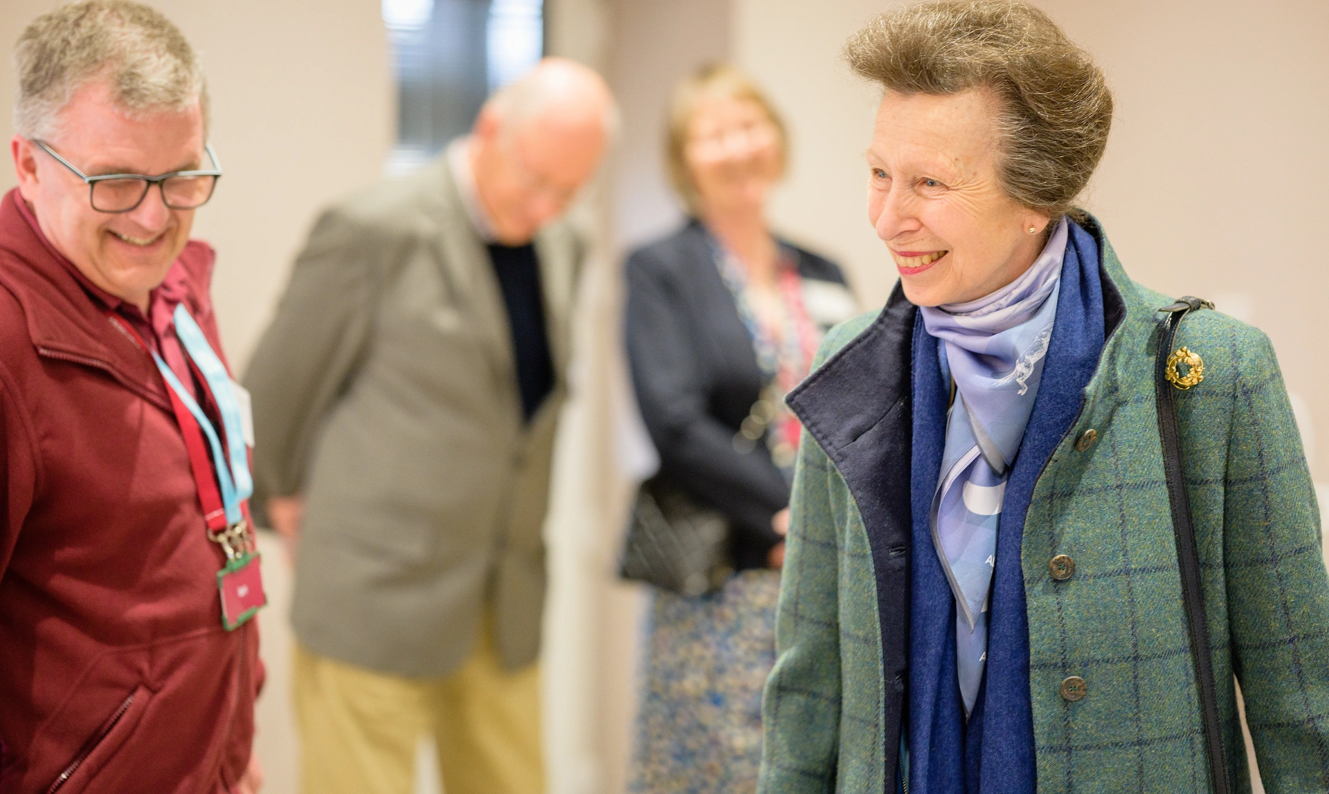  HRH The Princess Royal in a green coat and blue scarf and blouse looking to the left and smiling with a Hearing Dogs volunteer and some people in the background
