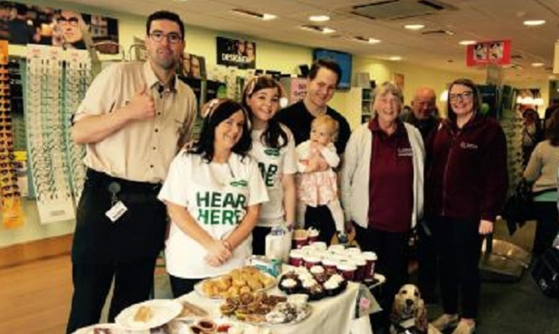Group of people standing by table full of cakes and coffee, smiling inside of a Specsavers store