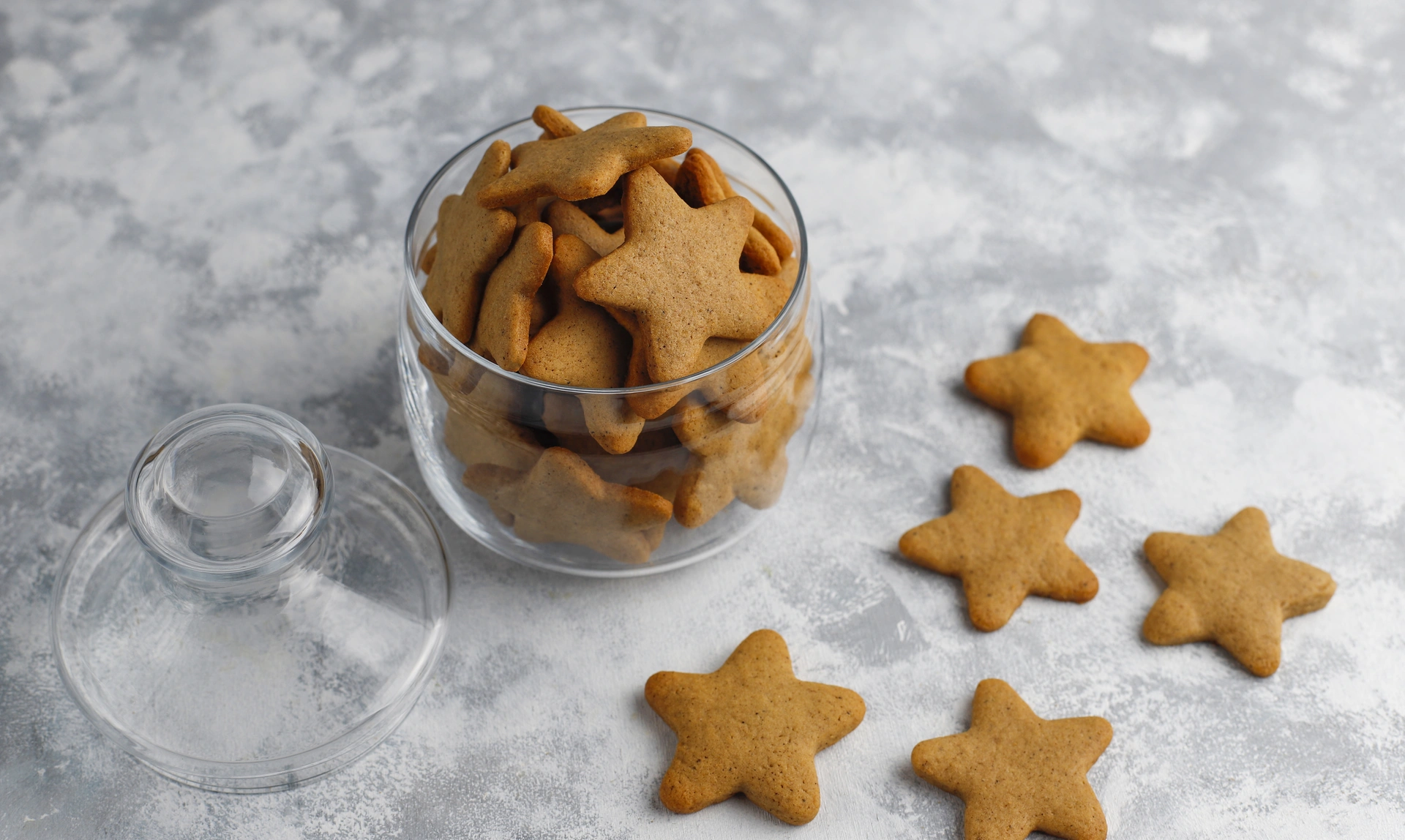 A glass jar filled with star-shaped gingerbread dog biscuits is open on a textured grey surface, with several biscuits scattered around it.