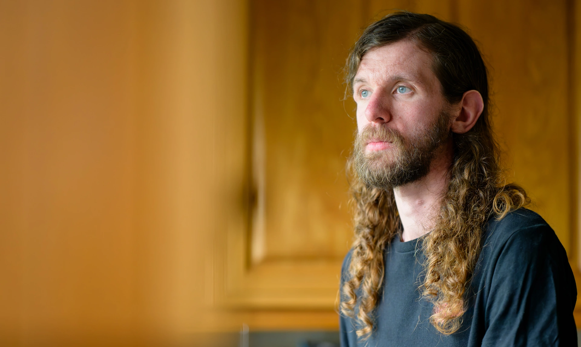 Head and shoulders image of a young man with long brown hair looking into the distance with a sad expression