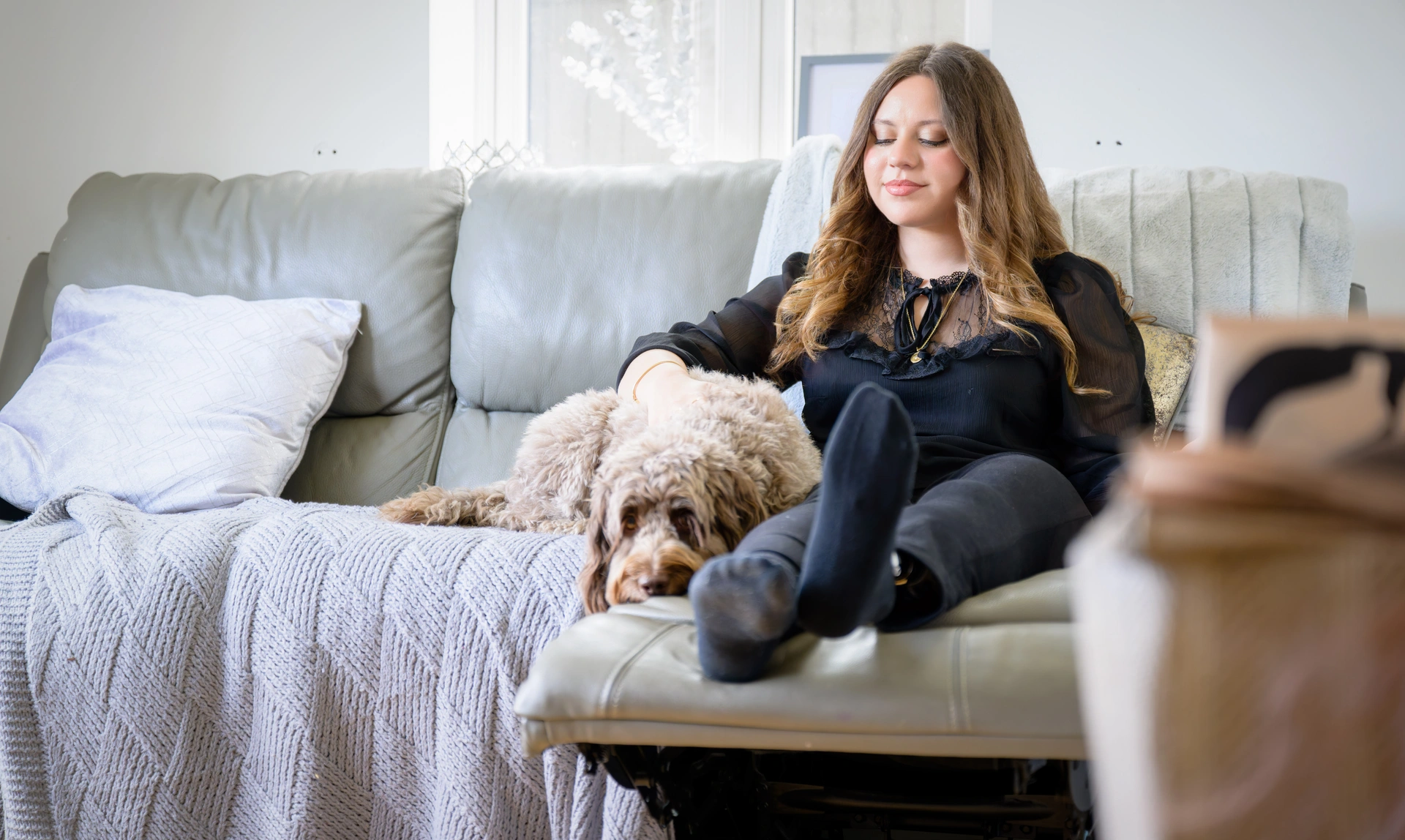  A young woman sat on a sofa smiling with her arm around a light brown cockapoo