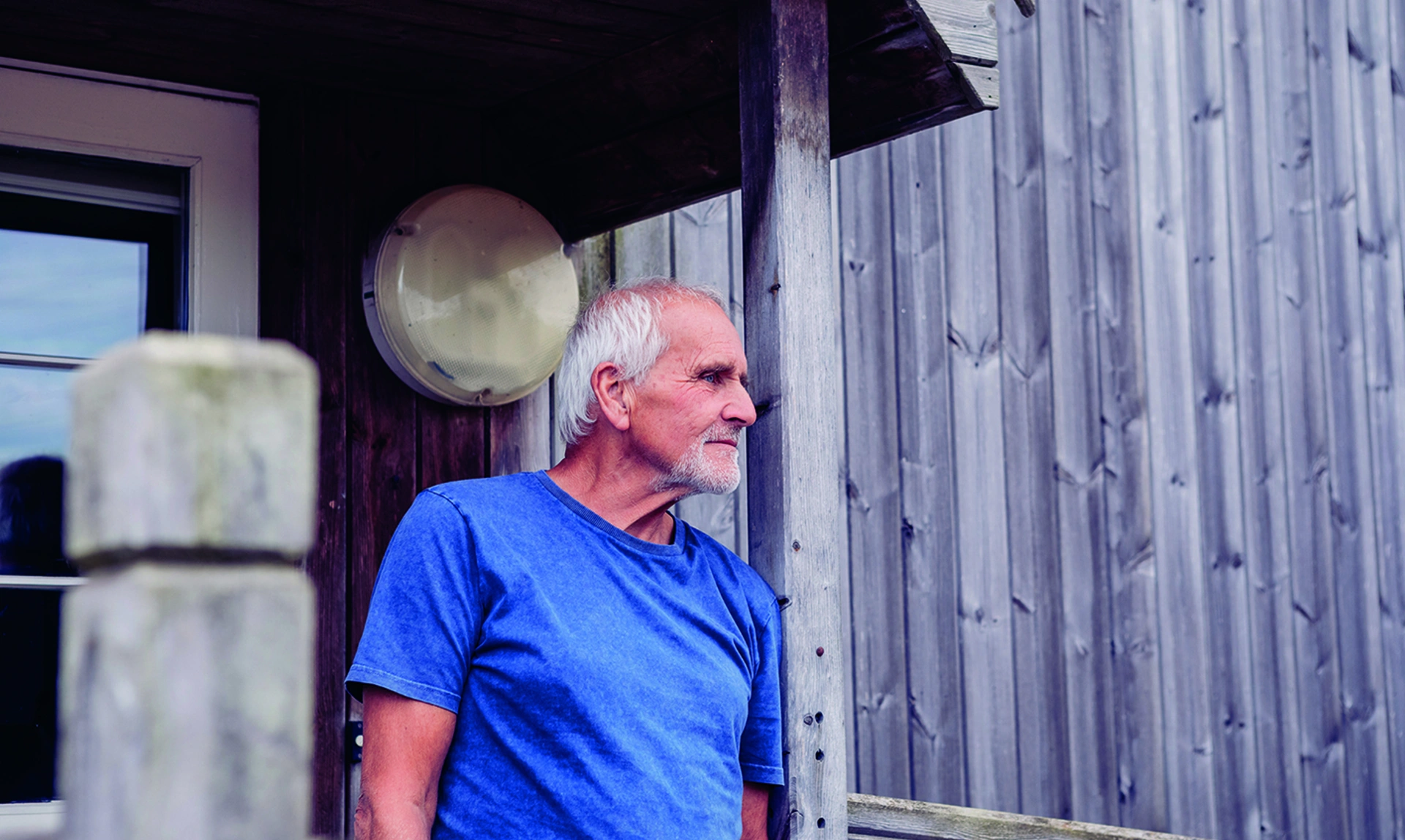 A man with a thoughtful expression stood on the porch of a wooden building looking into the distance, he has no visible signs of hearing loss