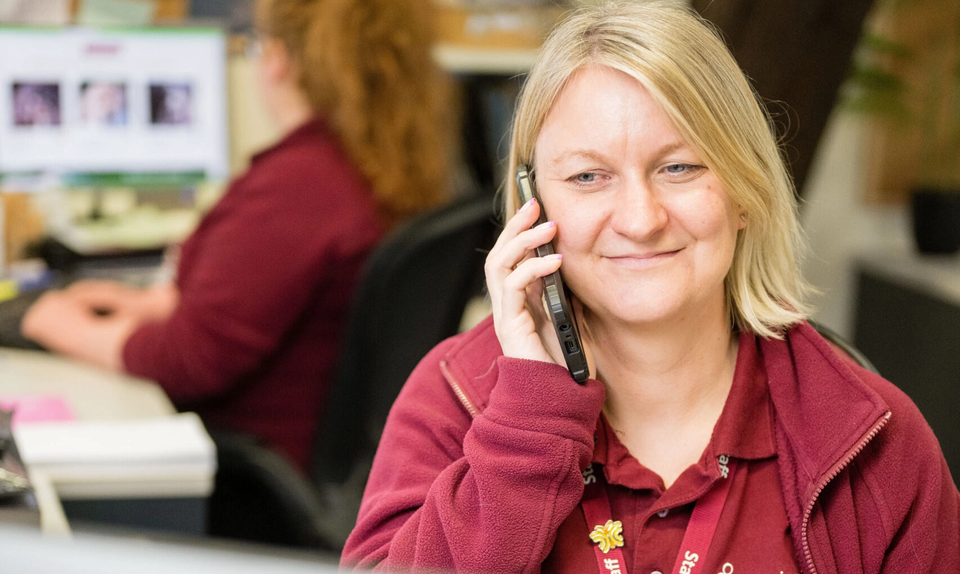 Woman, smiling, in hearing dogs uniform sat in an office in front of a laptop on the telephone