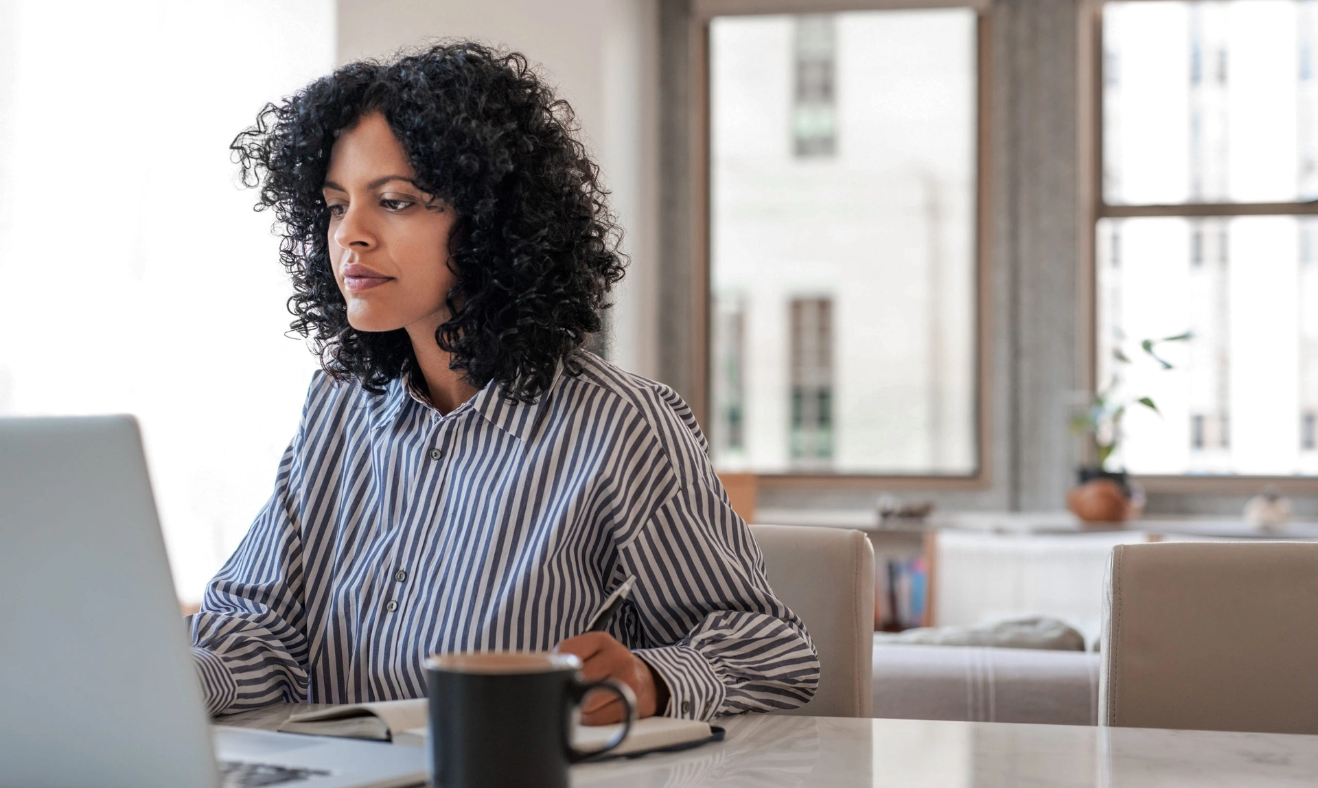 A woman sat working at a laptop, she appears to be reading the screen and making some notes in a notepad
