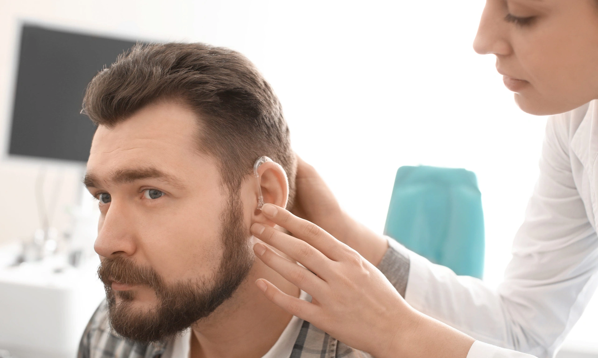 An audiologist leaning over and fitting a hearing aid to a man who is sat down