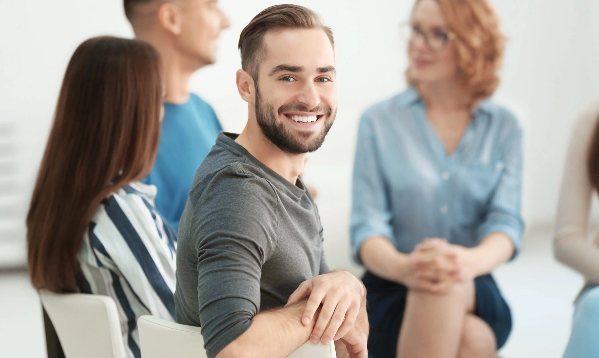 Smiling man sat in a chair turning around to look at the camera with other people sat talking in the background