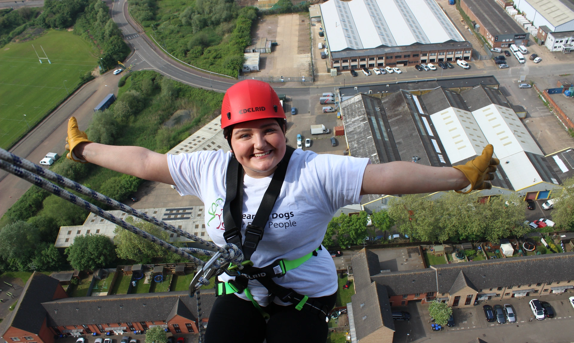 A person in a hard hat and harness hanging far above streets and fields looking happy with their arms outstretched 