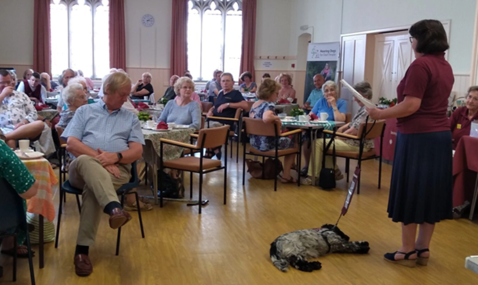 Speaker standing giving a talk to seated people in a hall with hearing dog lying down near her.