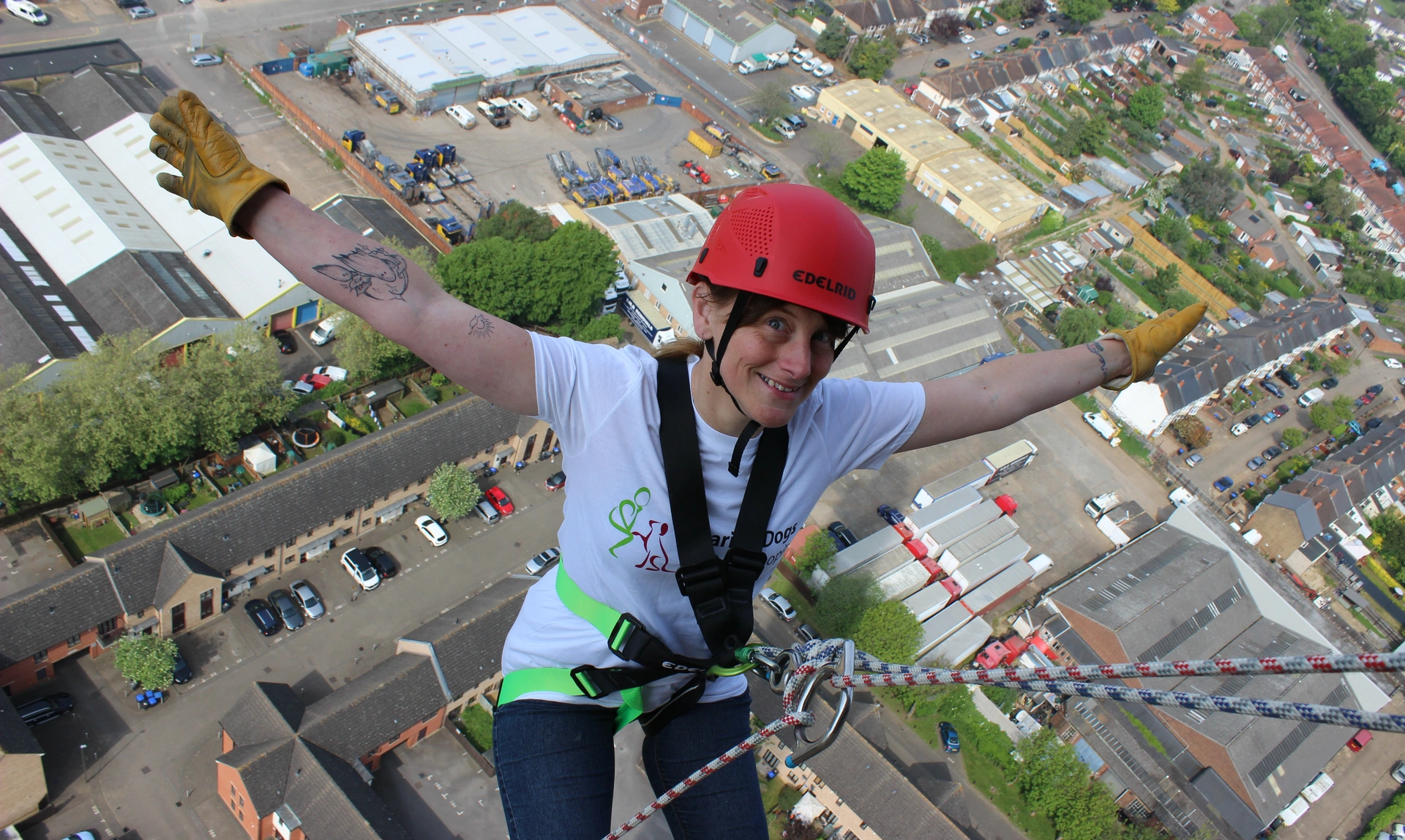 A woman, in a hard hat and harness, with her arms outstretched, looking happy while she abseils, with the ground far below her