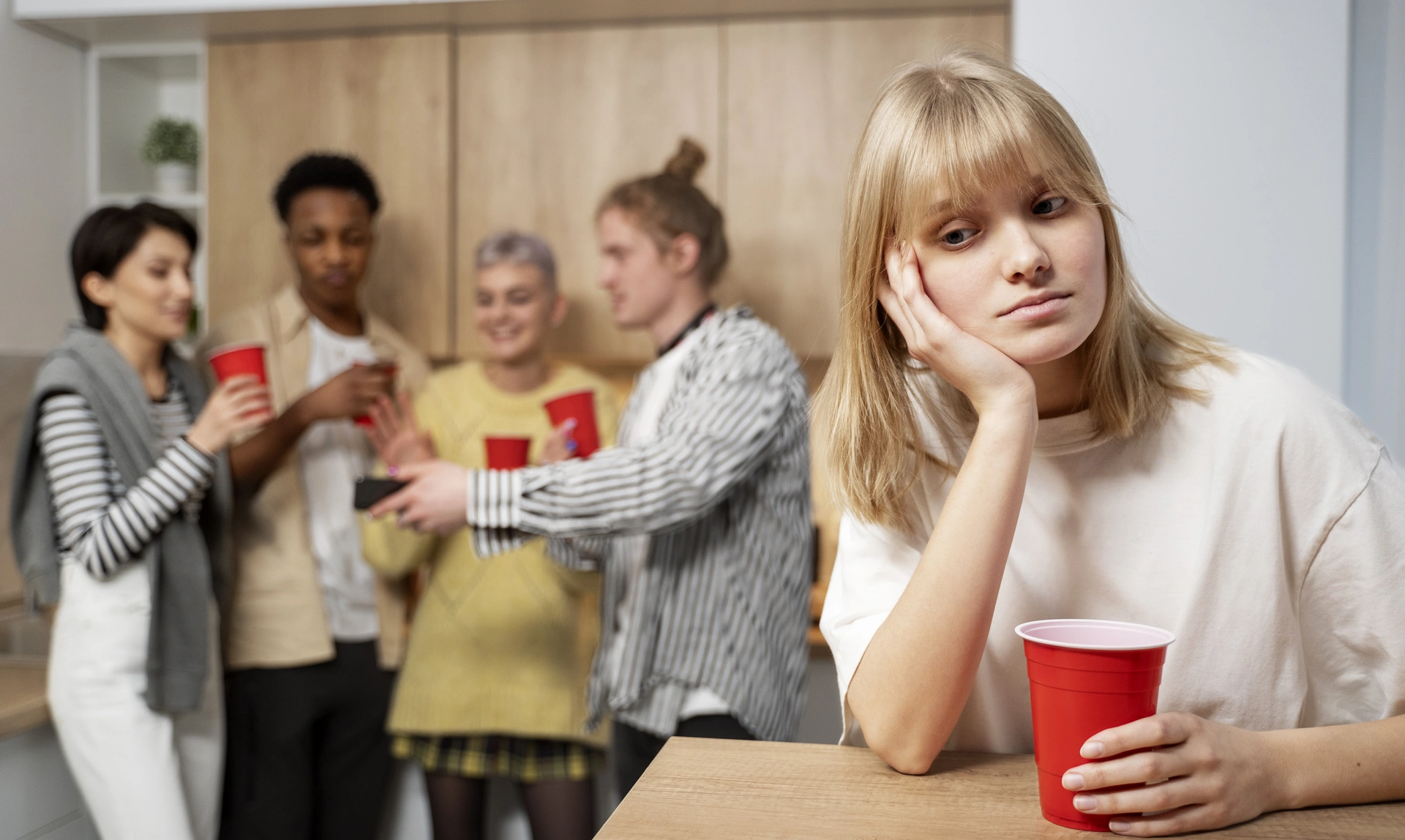 A woman sits alone at a table holding a paper cup and looing sad while 4 people are stood chatting and happy in the background