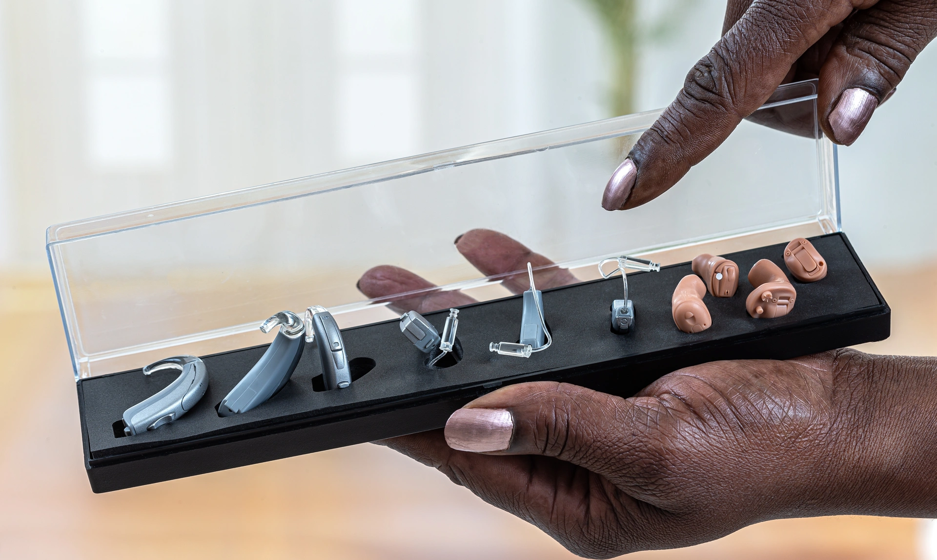 Close up of one hand holding a display case of four different types of hearing aid while the other hand points at them