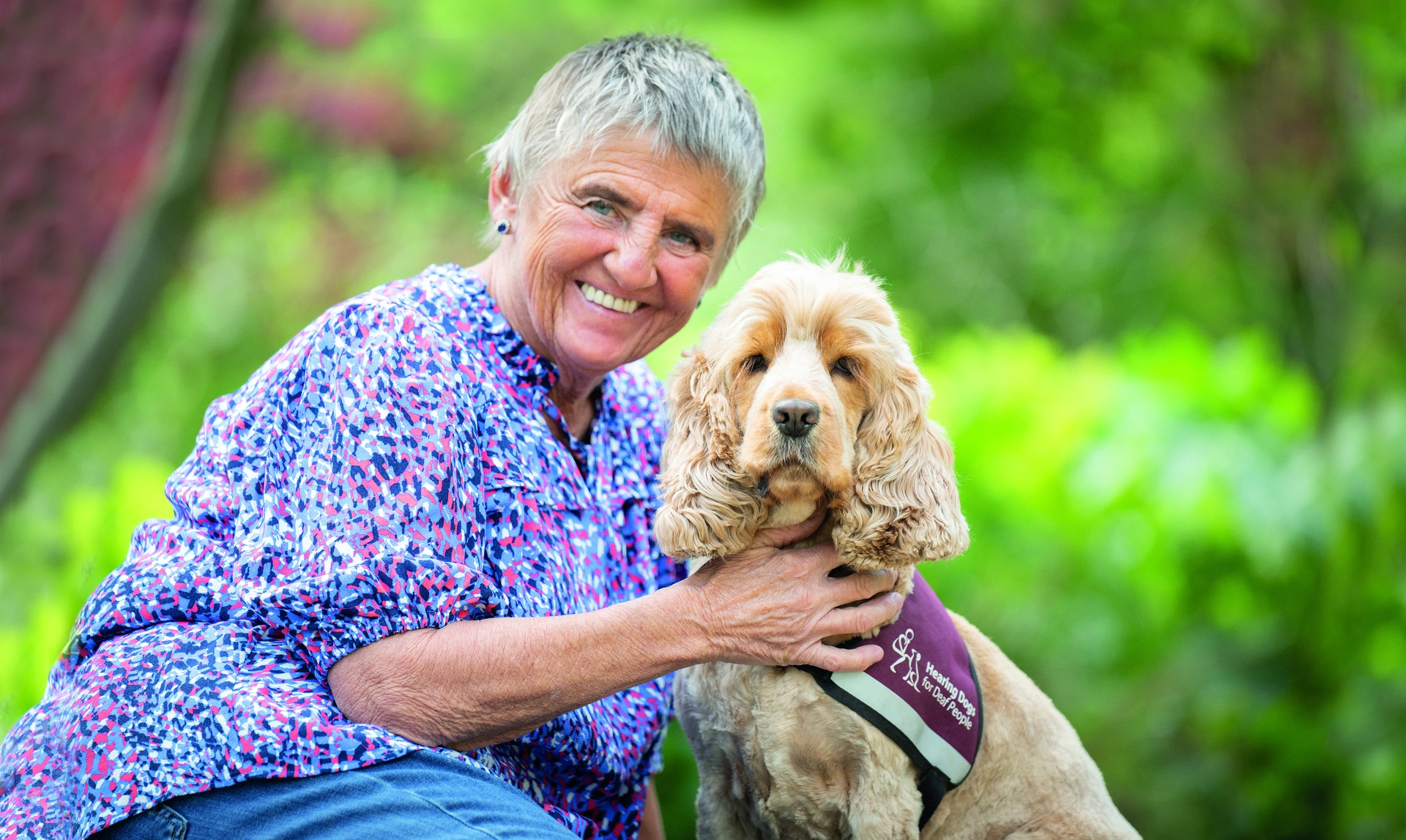  A woman sat with her hearing dog outside looking happy with greenery in the background