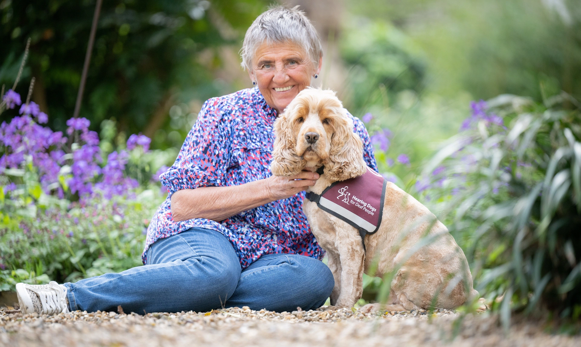 A smiling woman sat on gravel out side surrounded by flowers with her Spaniel hearing dog sat next to her 