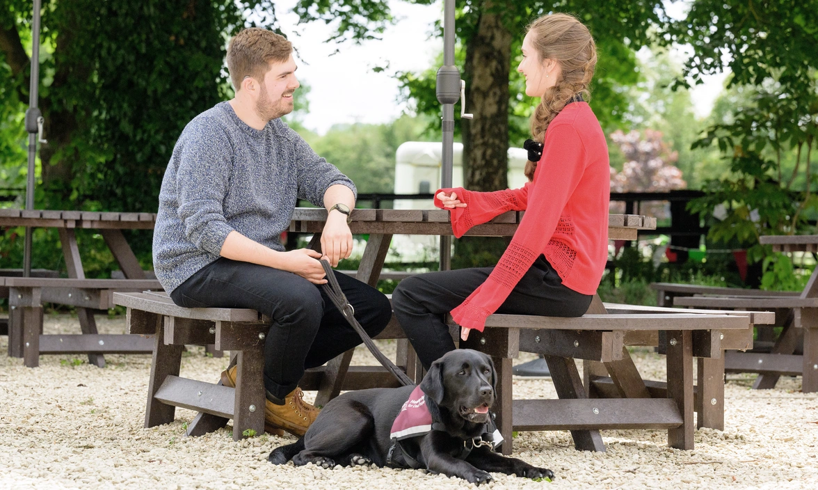 Two people sat on a bench outside chatting, one of them had a hearing dog wearing a qualified jacket
