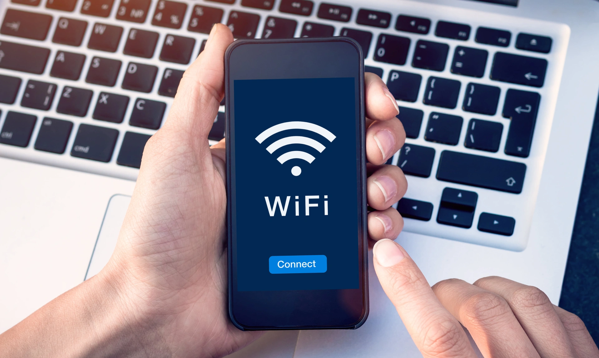 A hand holding a phone over the top of a laptop keyboard, with the wifi symbol and a 'Connect' button on the phone screen