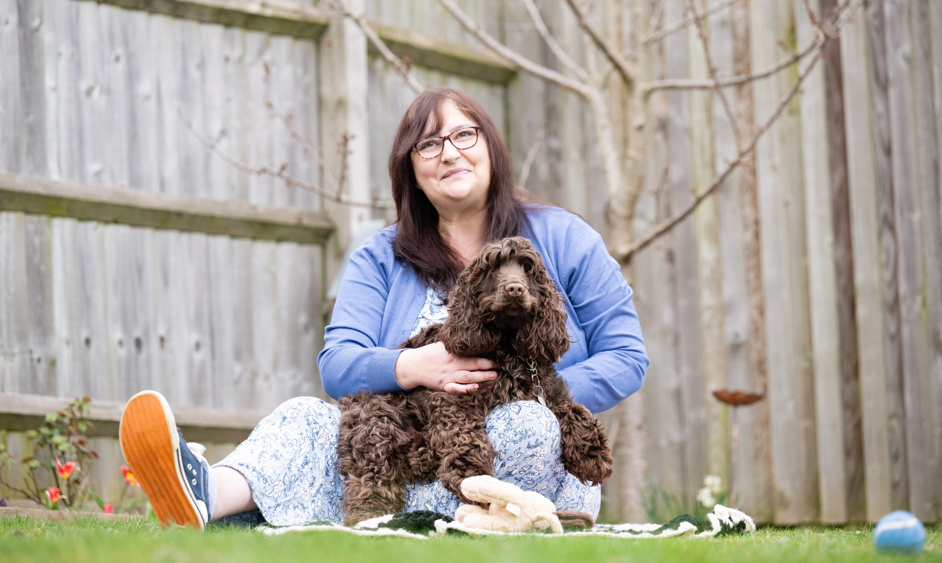 A bespectacled woman wearing a blue cardigan over a long dress is sitting on grass holding a brown curly haired dog in her arms. 