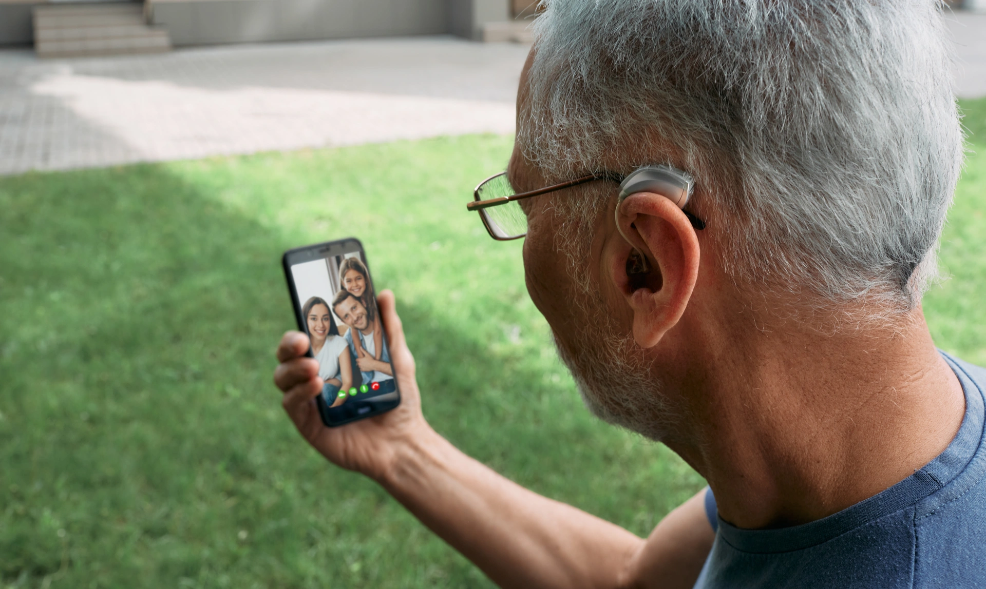 Older man wearing a hearing aid and holding his phone out in front of him that shows he is on a video call with a group of people who all look happy