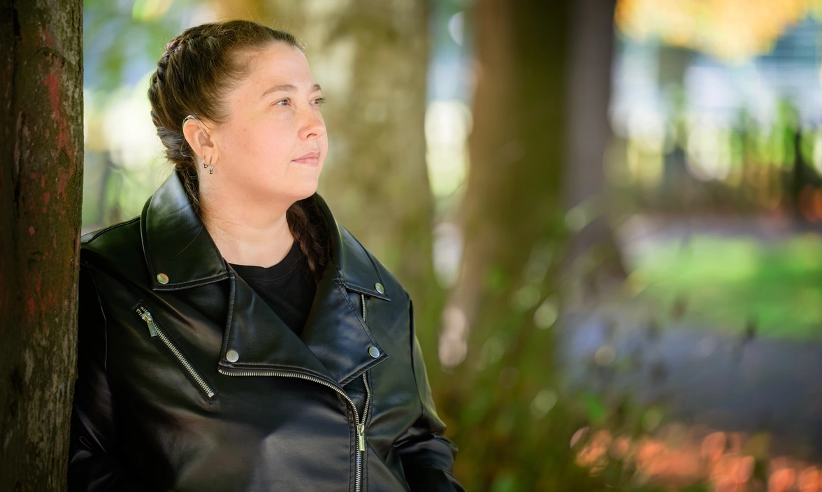 A woman in a black leather jacket leaning against a tree and looking off to the right. Blurred trees can be seen in the background.
