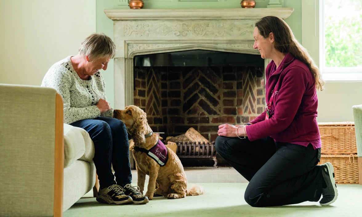 A hearing dogs staff member crouching in front of a woman with a hearing dog sat on a sofa