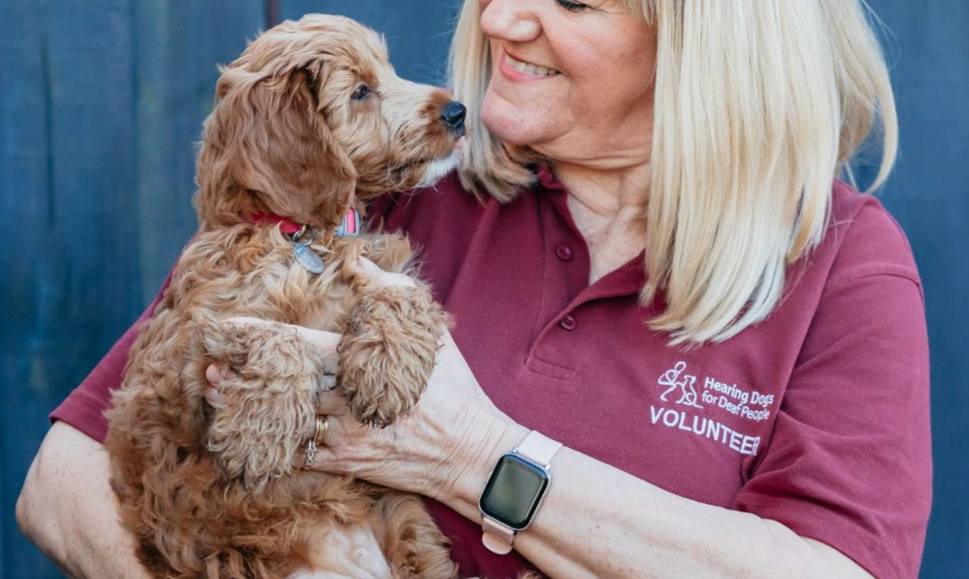 A blonde woman wearing a hearing dogs volunteer tshirt is standing smiling while holding a small puppy