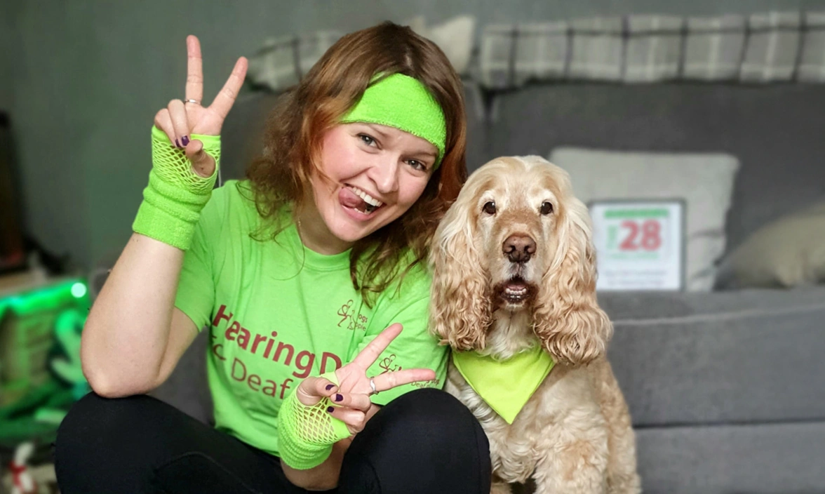 A woman in a sweatband and leg warmers smiling and sticking out her tongue, sat next to a dog smiling 