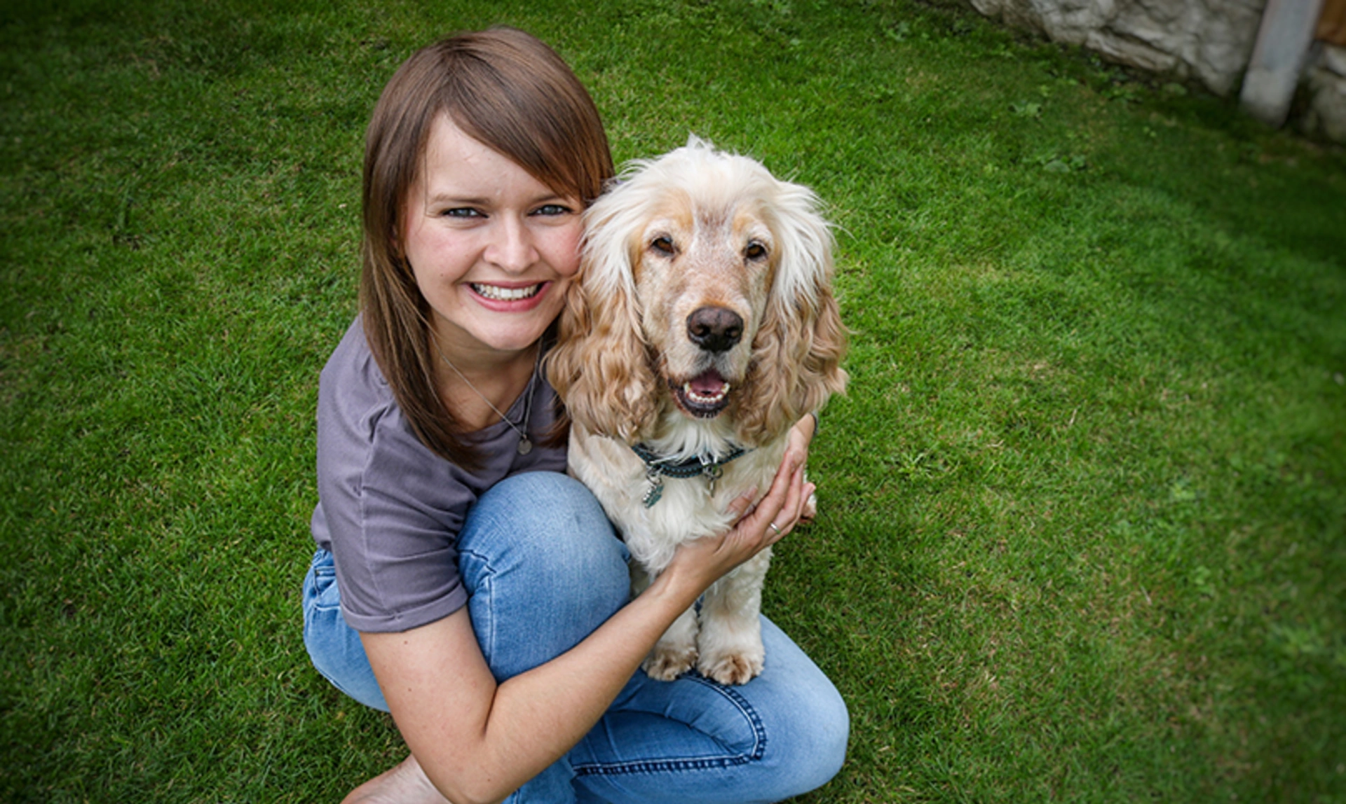 A young woman and light coloured dog are sitting on grass. The woman is smiling and hugging the dog.