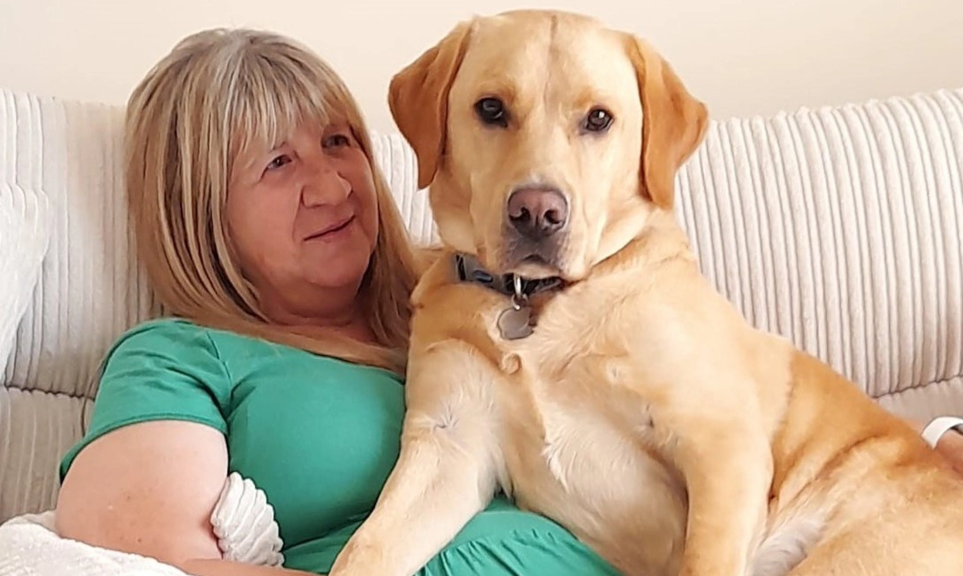 A woman with shoulder length blonde hair, sitting on a white sofa with a yellow labrador beside her