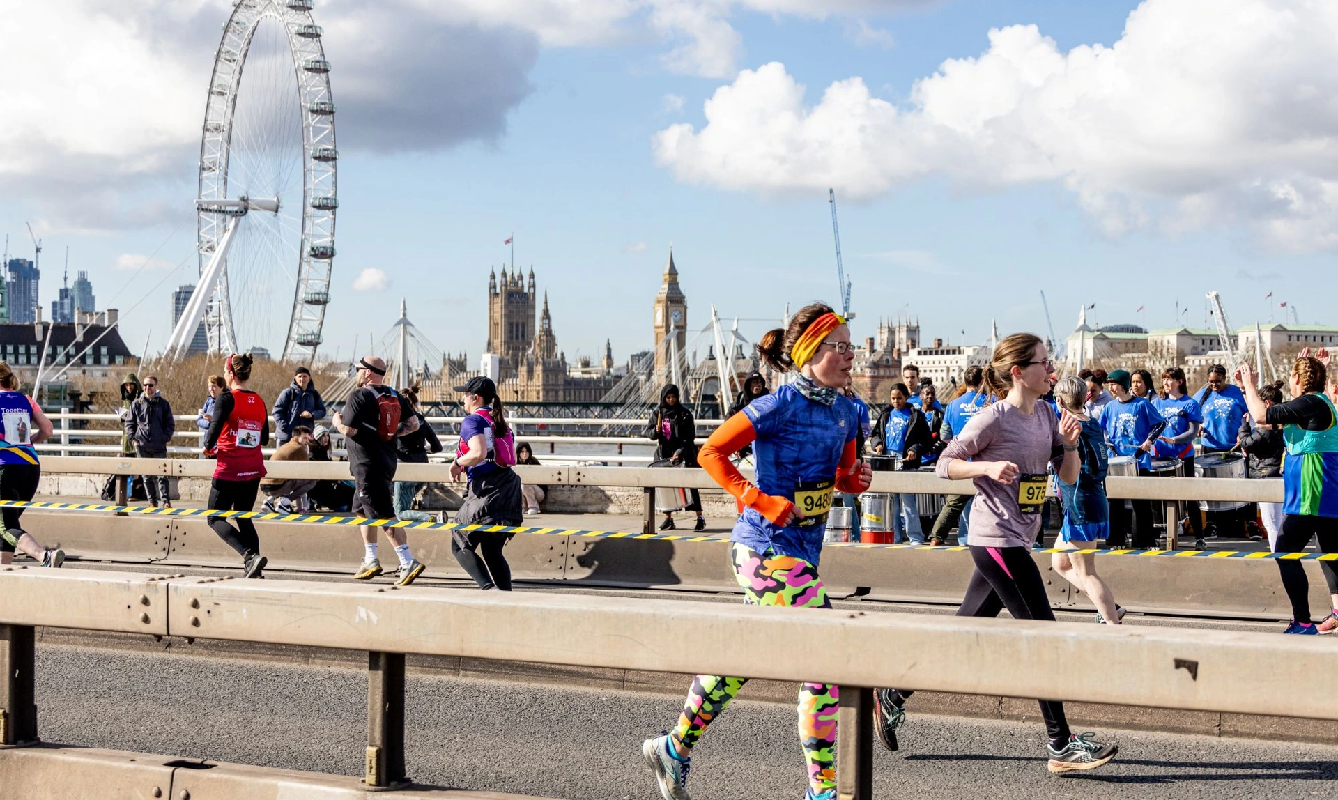 A group of marathon runners on a road. The London Eye can be seen in the background, as well as Big Ben and Westminster