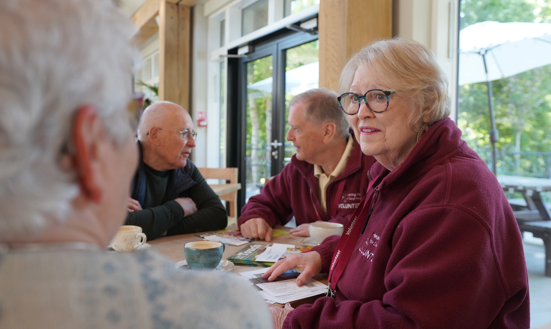 Seen over a woman's shoulder, a man and woman wearing hearing dogs volunteer uniforms are chatting to the other people at a table.