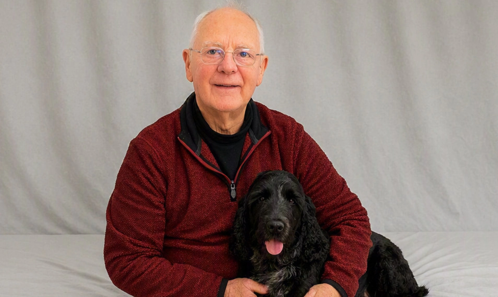 A smiling older gentleman sitting with a black dog on his lap, one arm around it. They are in a room draped with white sheets.