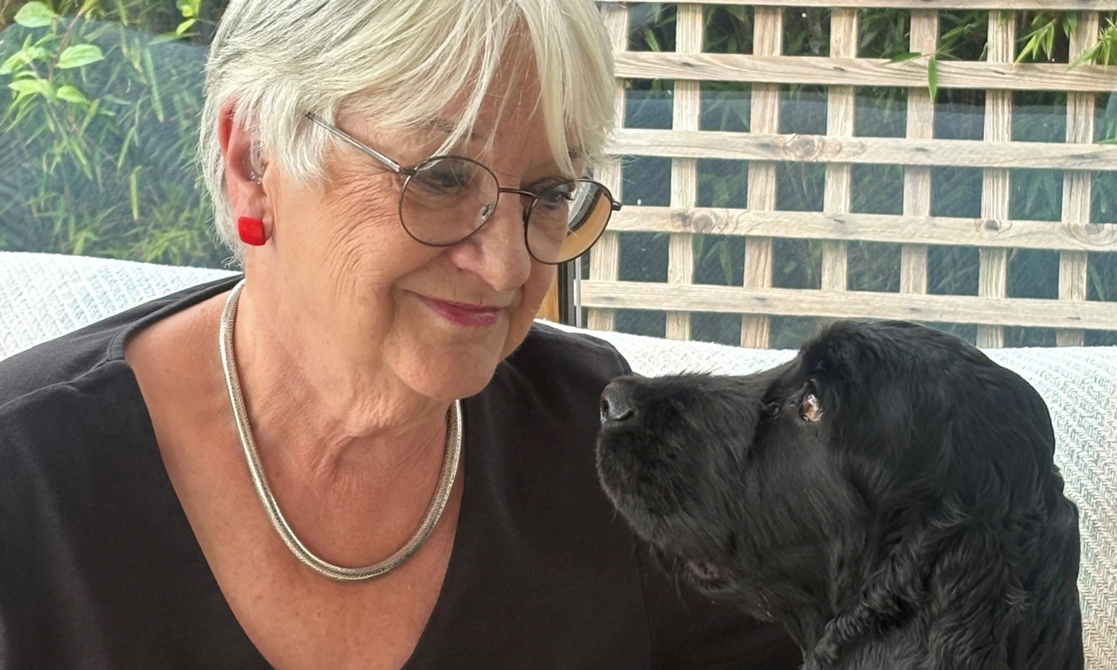 A bespectacled woman with white hair smiling at a black cocker spaniel sitting on a sofa beside her
