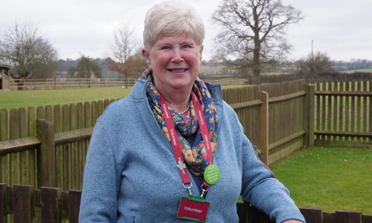 Mid shot of a happy woman leaving on a fence outside looking directly at the camera and wearing a lanyard with a badge that says ‘Volunteer’