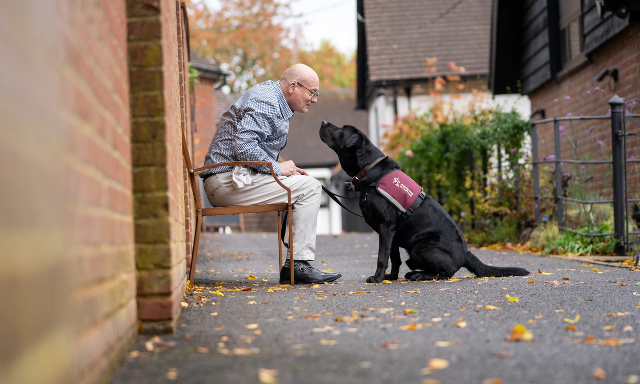 A profile photo of a man and a black labrador hearing dog facing each other. The man is sitting on a bench next to a wall.